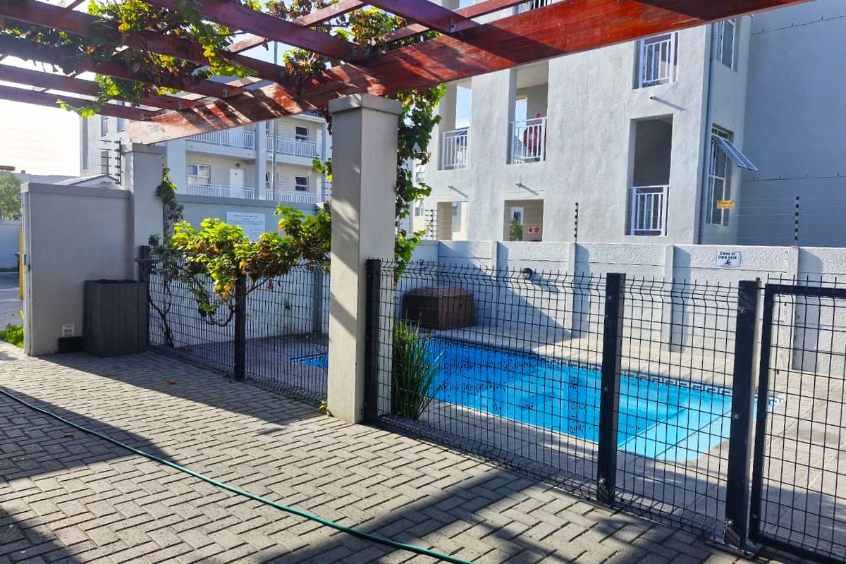 A communal outdoor pool area featuring a paved patio, a timber pergola structure, and secure perimeter fencing within a residential flat complex.