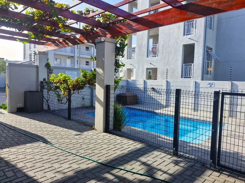 A communal outdoor pool area featuring a paved patio, a timber pergola structure, and secure perimeter fencing within a residential flat complex.