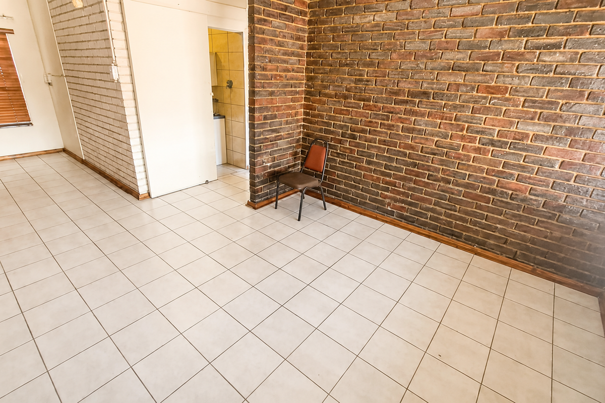 Spacious living area featuring a prominent exposed brick wall, neutral ceramic floor tiles, and natural light through venetian blinds.