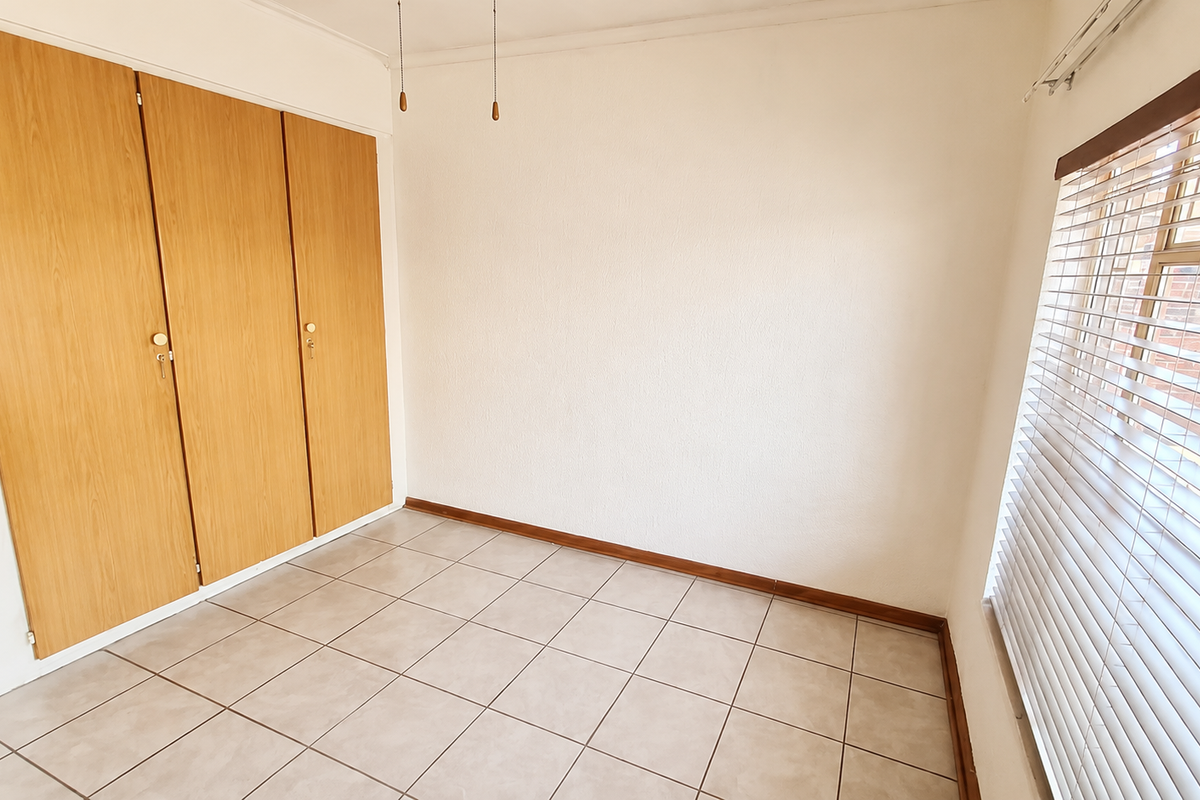 A bright, empty bedroom featuring neutral tiled flooring, built-in wooden cupboards, and fitted venetian blinds.