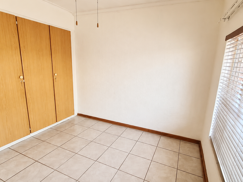A bright, empty bedroom featuring neutral tiled flooring, built-in wooden cupboards, and fitted venetian blinds.