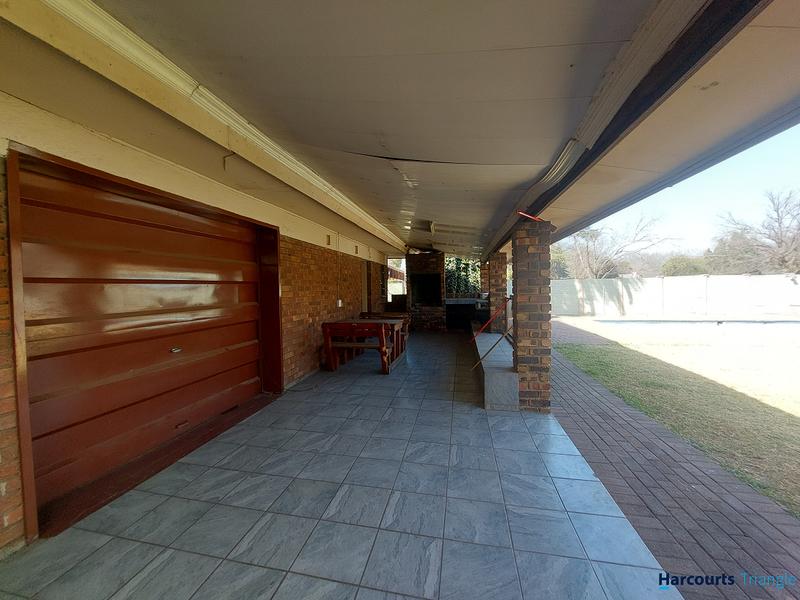Covered outdoor patio area featuring a built-in braai, tiled flooring, and brick pillars, overlooking the garden and pool area.