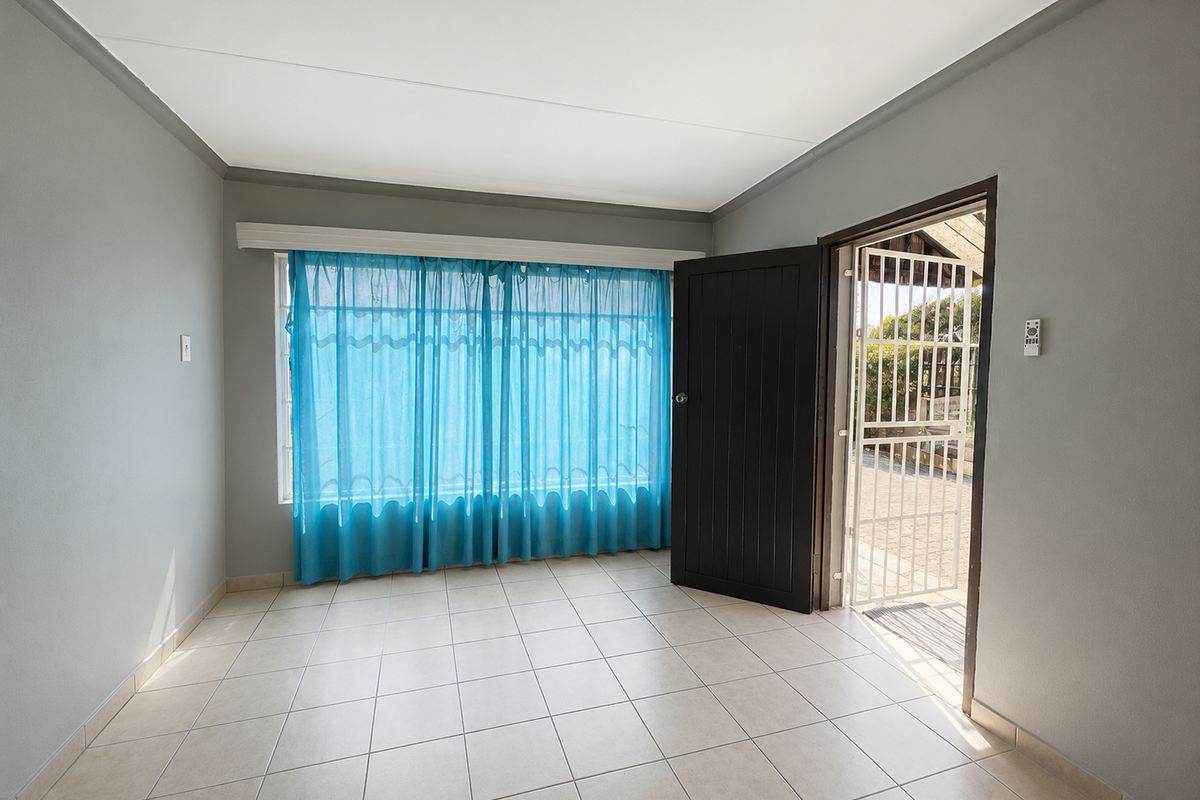A bright, empty living room featuring neutral tiled flooring, clean grey walls, and direct access to an outdoor area through a secure door.
