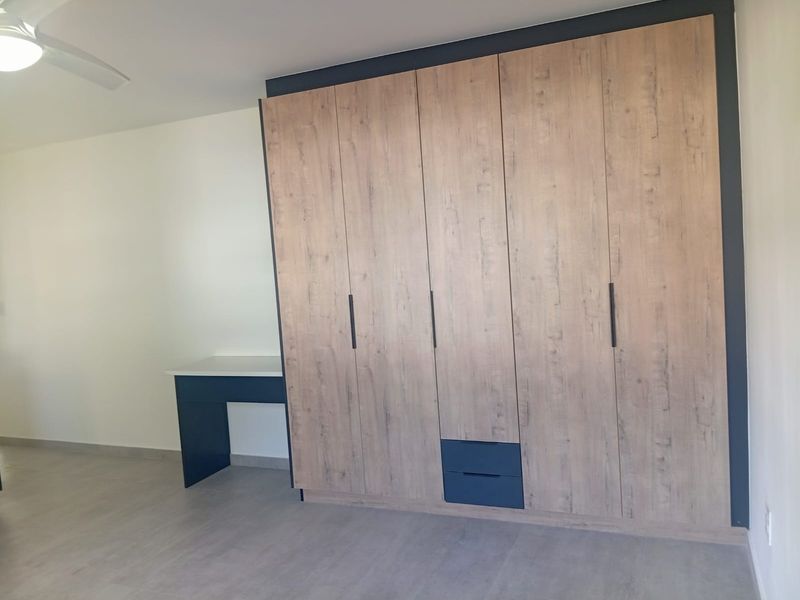 A clean, modern bedroom featuring a large built-in wardrobe with wood-grain finish, integrated desk space, and a ceiling fan.