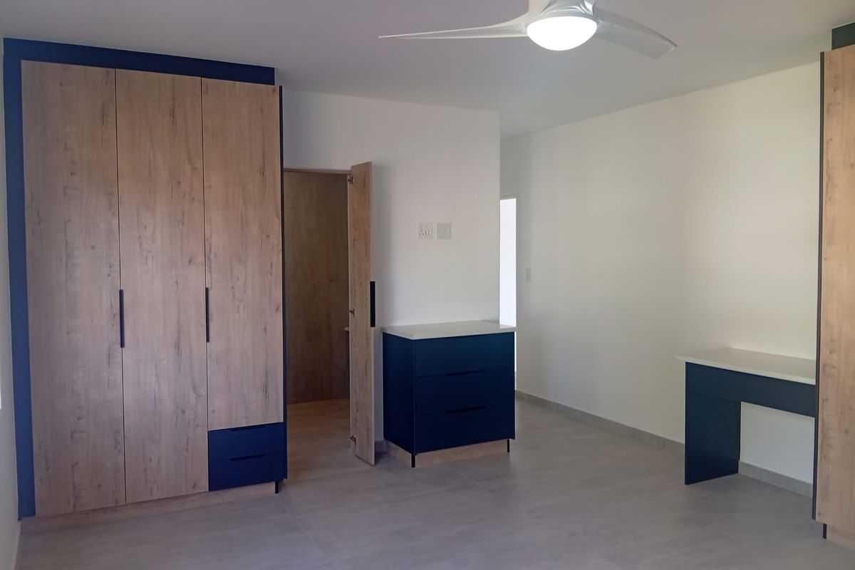 A modern, spacious bedroom featuring built-in wooden cupboards, a matching built-in desk, tiled flooring, and a contemporary ceiling fan.