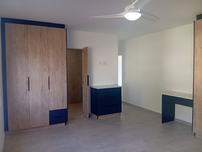 A modern, spacious bedroom featuring built-in wooden cupboards, a matching built-in desk, tiled flooring, and a contemporary ceiling fan.