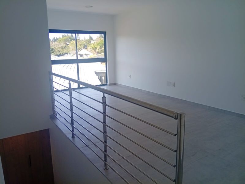 A bright, empty upper-level living area featuring modern tiled flooring, a sleek stainless steel balustrade, and large windows with neighborhood views.