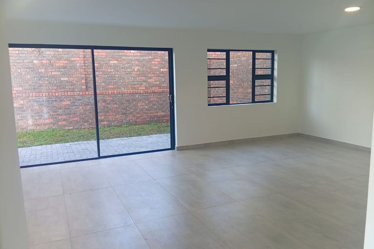 A bright, empty living area featuring large format floor tiles, modern aluminium window frames, and a sliding door leading to a paved outdoor space.