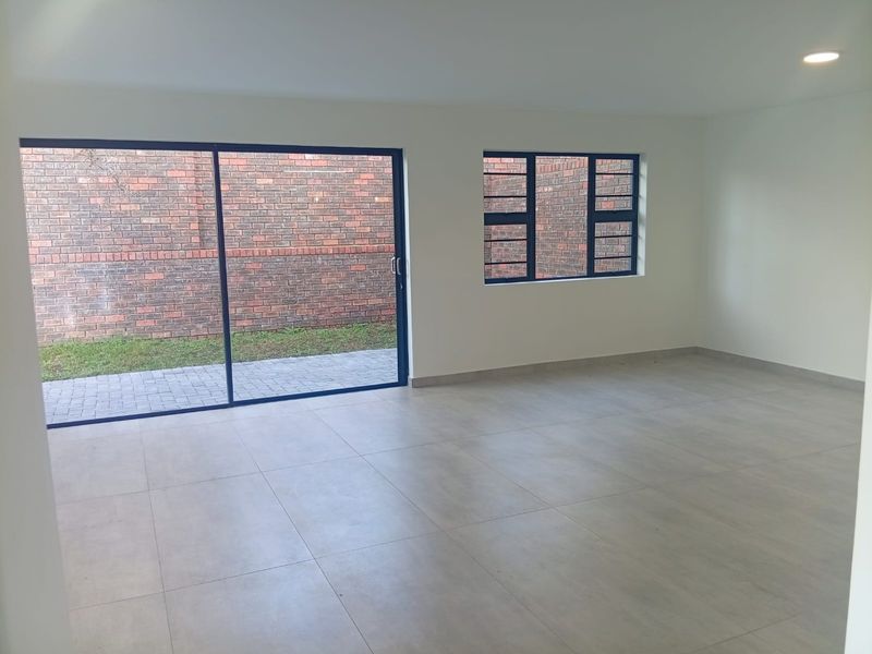 A bright, empty living area featuring large format floor tiles, modern aluminium window frames, and a sliding door leading to a paved outdoor space.