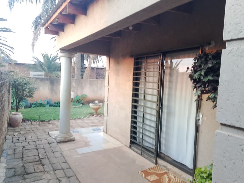 A covered patio area featuring a sliding glass door with security gate, concrete column, and paved flooring, overlooking a small garden space.