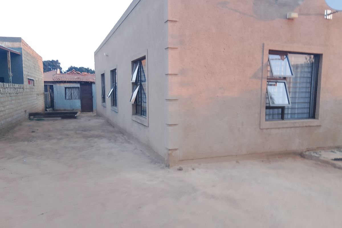 Side and rear view of a plastered masonry house featuring top-hung aluminium windows with security bars and an unpaved yard area.