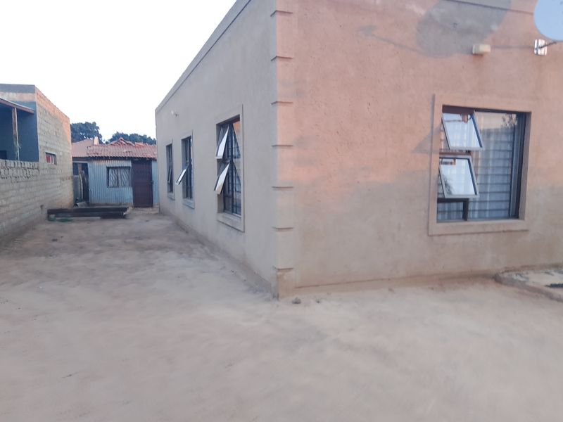 Side and rear view of a plastered masonry house featuring top-hung aluminium windows with security bars and an unpaved yard area.