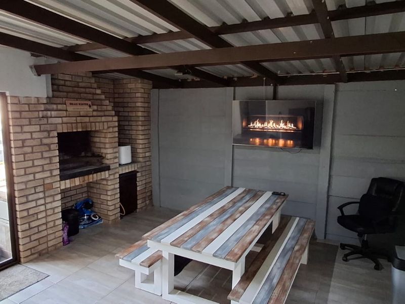 Covered outdoor patio featuring a built-in brick braai, wall-mounted electric fireplace, tiled flooring, and direct access to the garden via sliding glass doors.