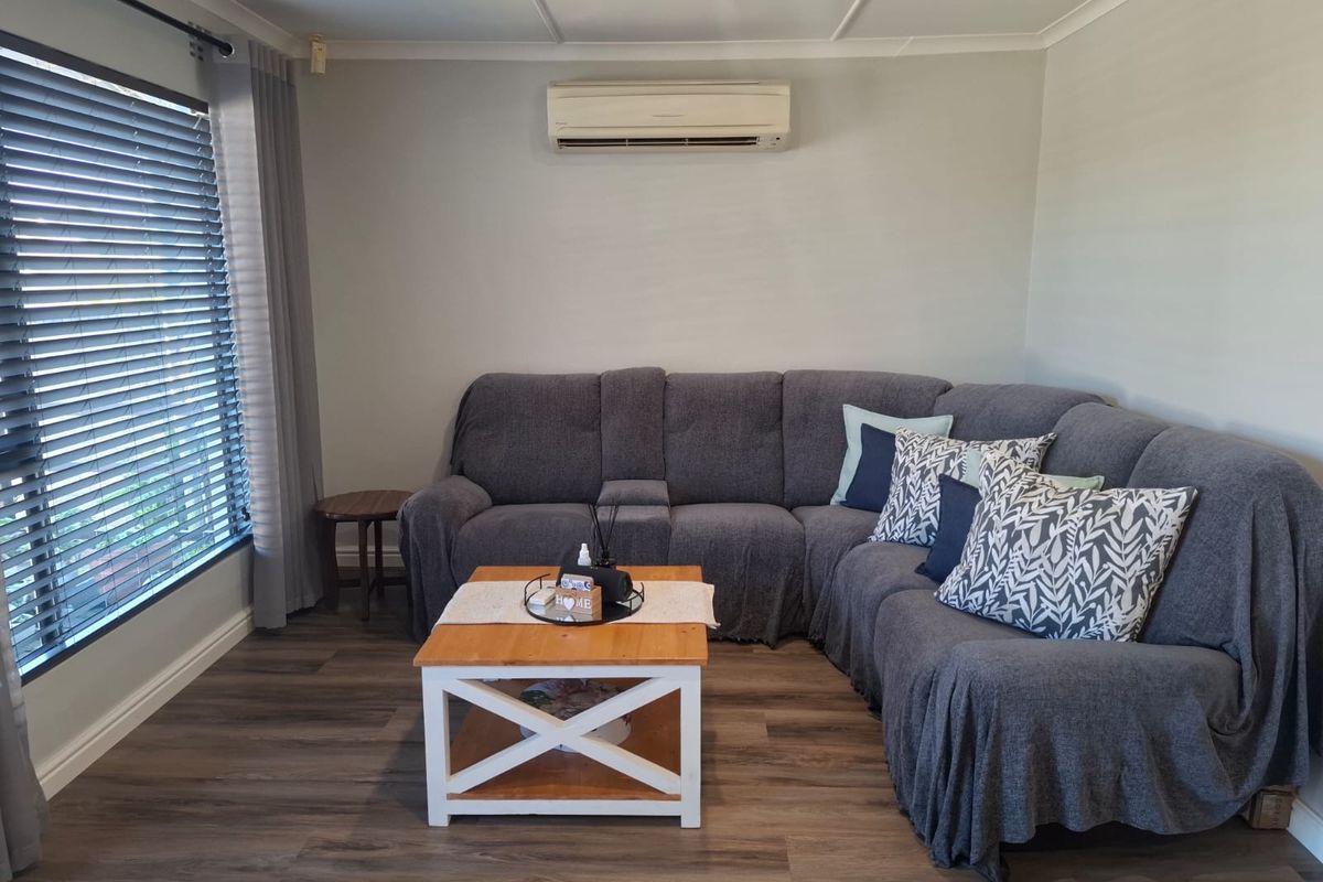 A comfortable living room featuring modern laminate flooring, a wall-mounted split-system air conditioner, and large windows fitted with venetian blinds.
