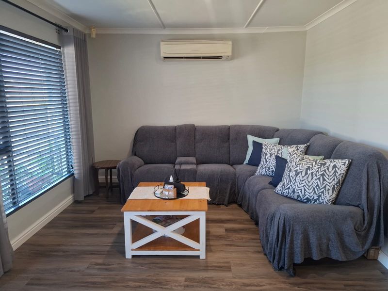 A comfortable living room featuring modern laminate flooring, a wall-mounted split-system air conditioner, and large windows fitted with venetian blinds.