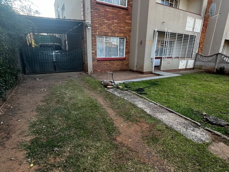 Front exterior view of the flat featuring a brick facade, carport, security gates, and a private garden area with a concrete walkway.