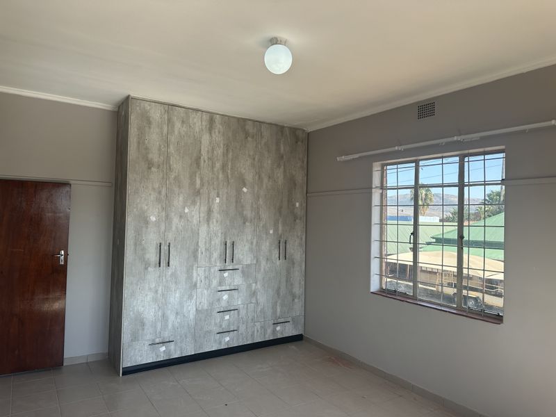 A spacious bedroom featuring neutral tiled flooring, built-in cupboards with a modern wood-look finish, and a window fitted with security bars.