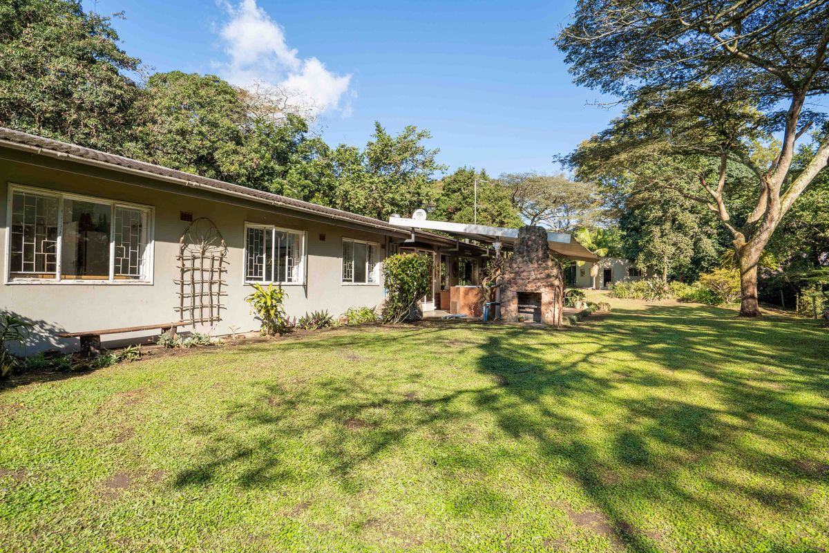 Rear view of the property featuring a spacious lawn, established trees, and a built-in stone fireplace on the covered patio area.
