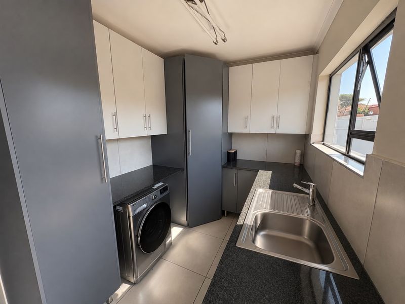 A modern scullery featuring sleek grey and white cabinetry, granite countertops, a stainless steel sink, and tiled flooring with natural light.