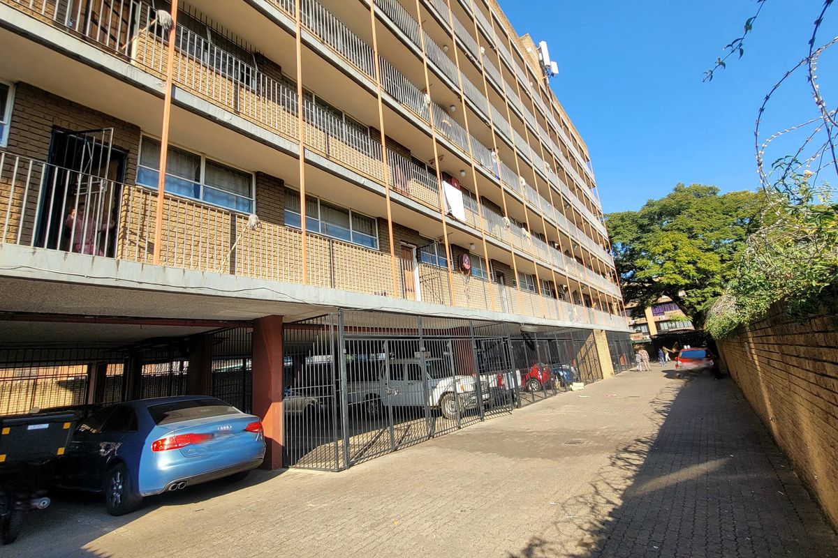 Exterior view of a multi-story apartment building featuring brick facade, private balconies with metal railings, and ground-level covered parking area.