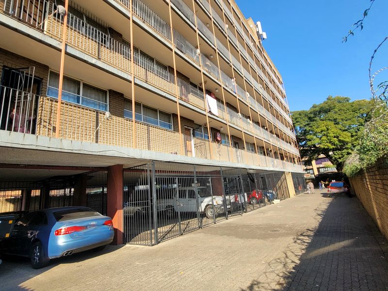 Exterior view of a multi-story apartment building featuring brick facade, private balconies with metal railings, and ground-level covered parking area.