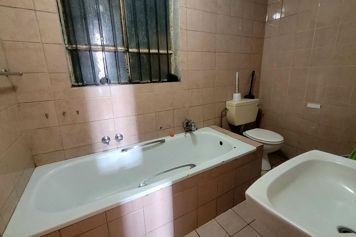A dated bathroom featuring a bathtub, toilet, and washbasin with full-height wall tiling and a window with security bars.