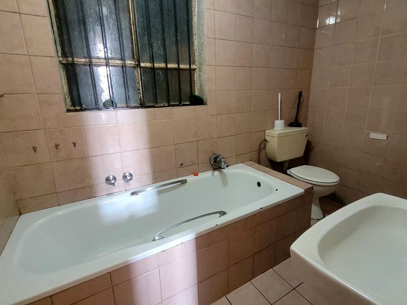 A dated bathroom featuring a bathtub, toilet, and washbasin with full-height wall tiling and a window with security bars.