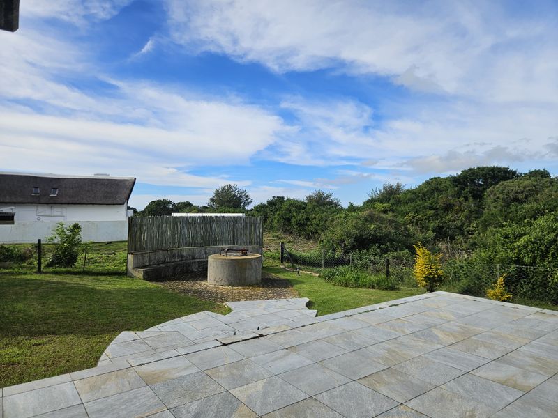 Outdoor patio area featuring a tiled floor, built-in braai, and concrete seating, overlooking a garden with natural vegetation and perimeter fencing.