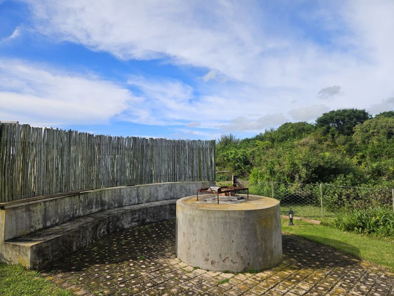 Outdoor entertainment area featuring a built-in concrete braai, curved bench seating, and paved flooring, enclosed by natural reed fencing.