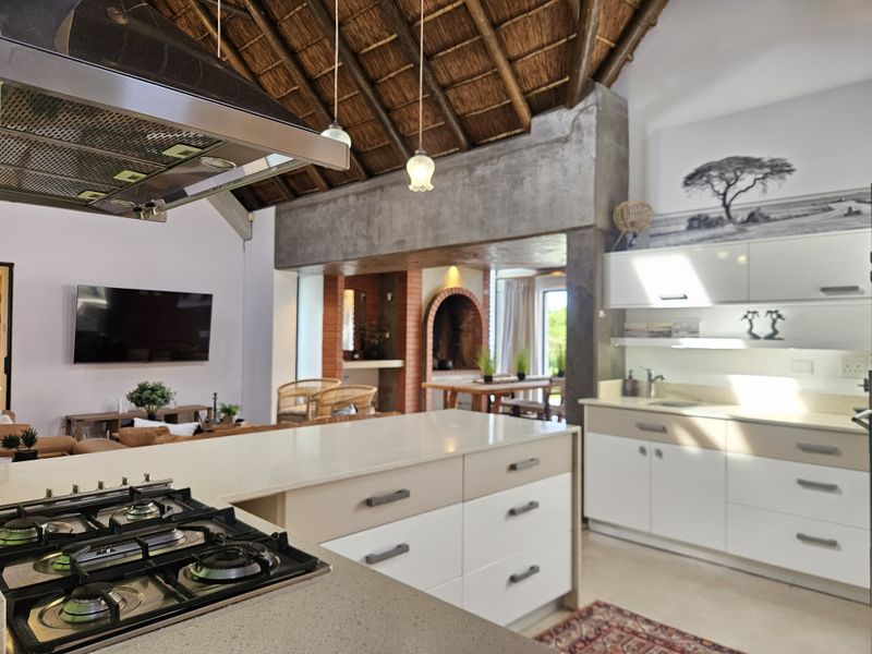 Spacious open-plan kitchen and living area featuring a gas hob, stone countertops, and a striking exposed thatch roof with a feature brick fireplace.