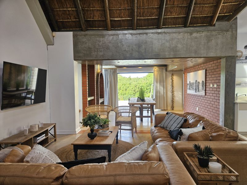Spacious open-plan living area featuring an exposed thatch roof, concrete structural beams, and exposed brickwork, leading out to a bright dining space.