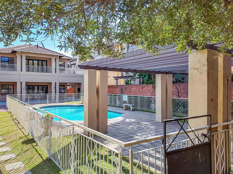Communal outdoor pool area featuring a paved patio, pergola structure, and secure metal fencing within an apartment complex setting.