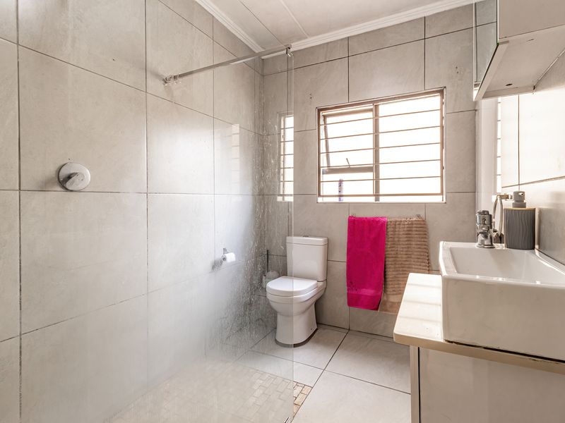 A functional bathroom featuring a glass-enclosed shower, tiled walls and floors, a vanity basin, and a toilet with natural light from a window.