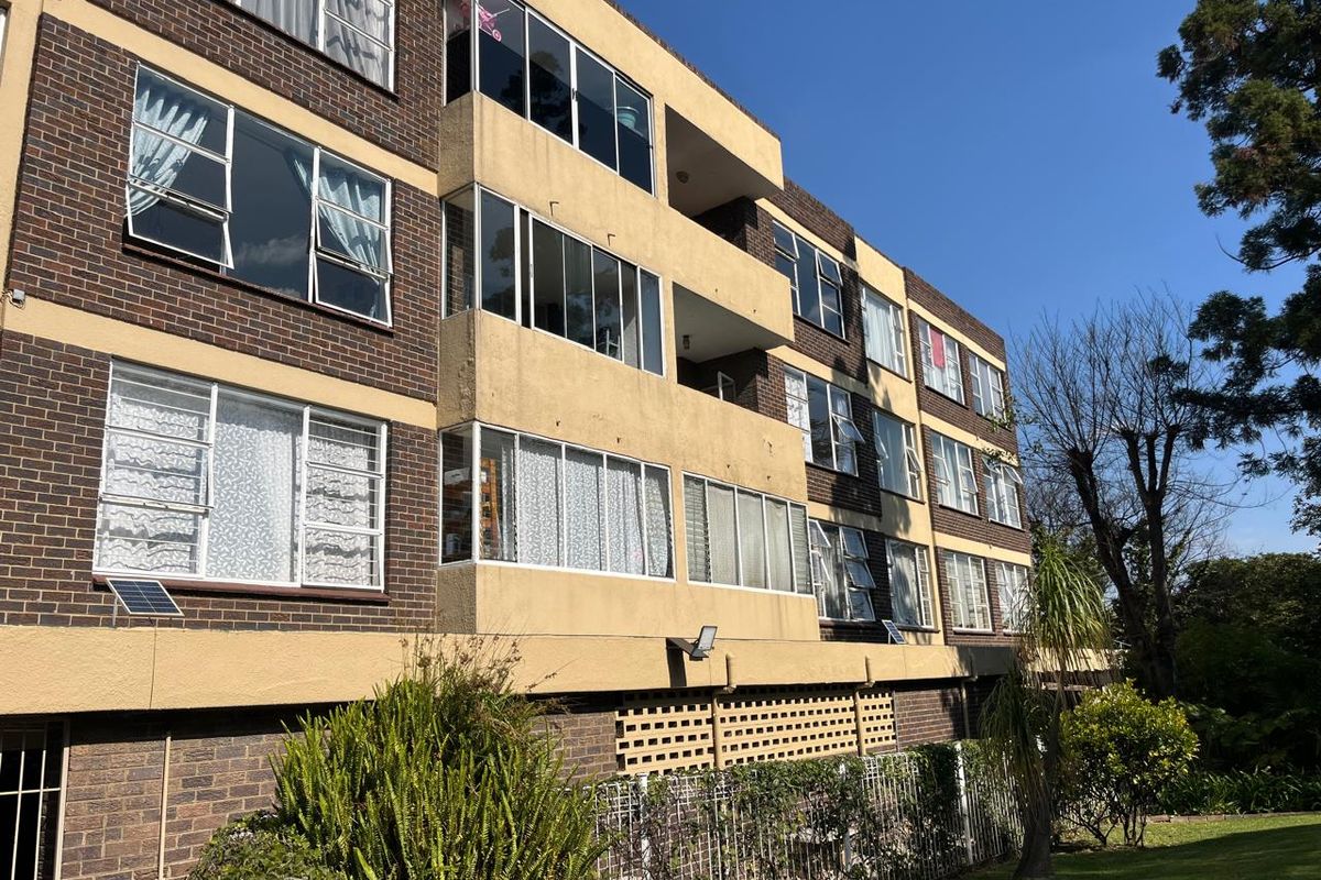 Rear elevation of a multi-story brick apartment building featuring enclosed balconies and security-fitted windows overlooking a landscaped garden area.