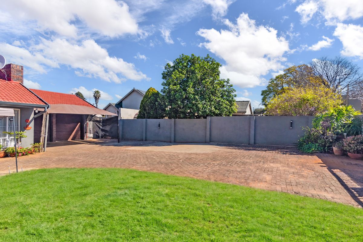 A spacious rear exterior area featuring a paved driveway, lawn, boundary wall, and access to a garage with an attached carport.