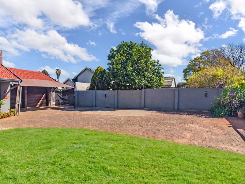A spacious rear exterior area featuring a paved driveway, lawn, boundary wall, and access to a garage with an attached carport.