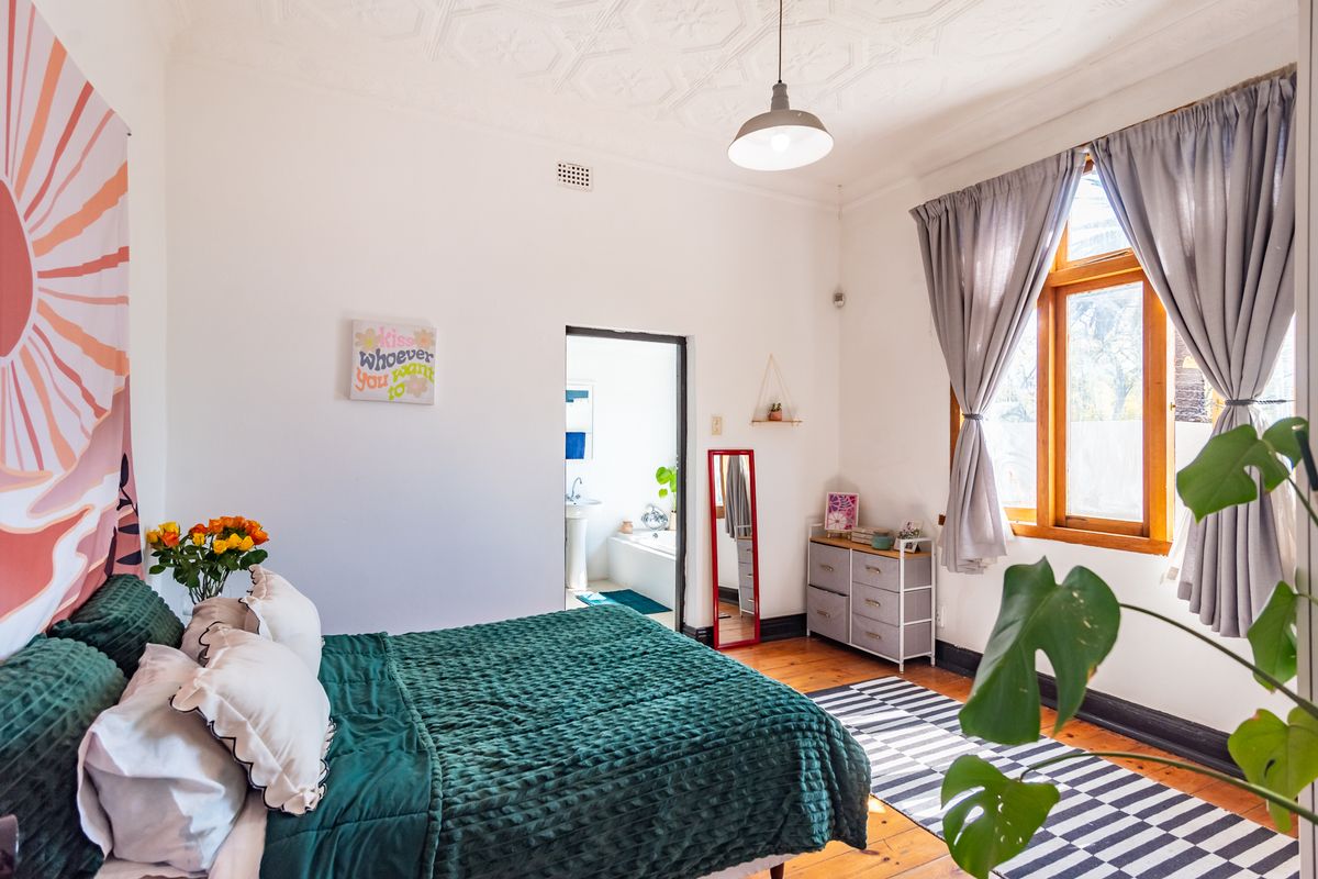 A bright bedroom featuring original timber flooring, a decorative pressed metal ceiling, and a window with timber frames, leading into an adjoining bathroom.