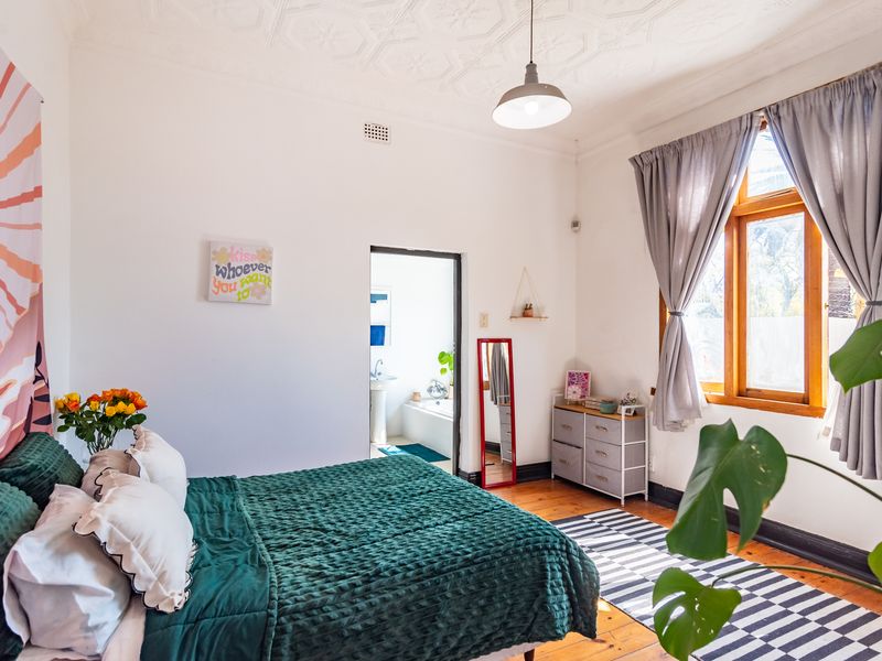 A bright bedroom featuring original timber flooring, a decorative pressed metal ceiling, and a window with timber frames, leading into an adjoining bathroom.