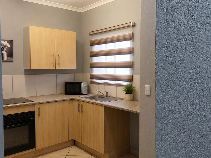 A compact, well-maintained kitchen featuring light wood-finish cabinetry, integrated oven and hob, and neutral tiled flooring with a functional layout.