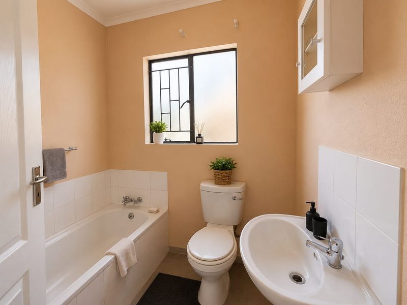 A functional bathroom featuring a bathtub, pedestal sink, and toilet, with neutral wall tones and a window fitted with security bars.