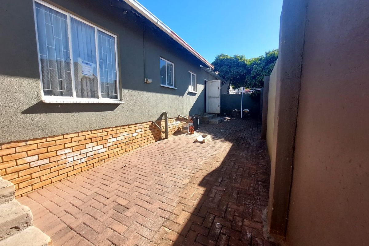 A paved rear exterior area featuring a brick-accented facade, security bars on windows, and a surrounding boundary wall.