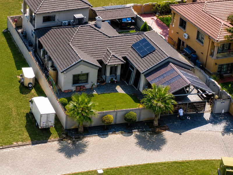Aerial view of a residential property featuring a tiled roof with solar panels, a covered patio, paved driveway, and well-maintained boundary walls.