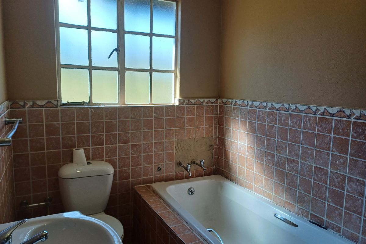 A dated bathroom featuring a built-in bathtub, pedestal basin, and toilet, with original tiled splashbacks and a steel-framed window.