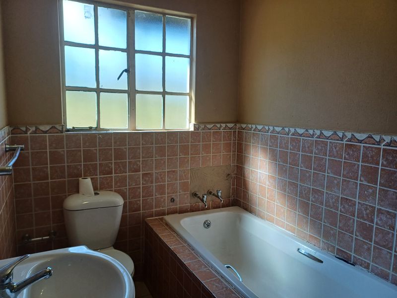 A dated bathroom featuring a built-in bathtub, pedestal basin, and toilet, with original tiled splashbacks and a steel-framed window.