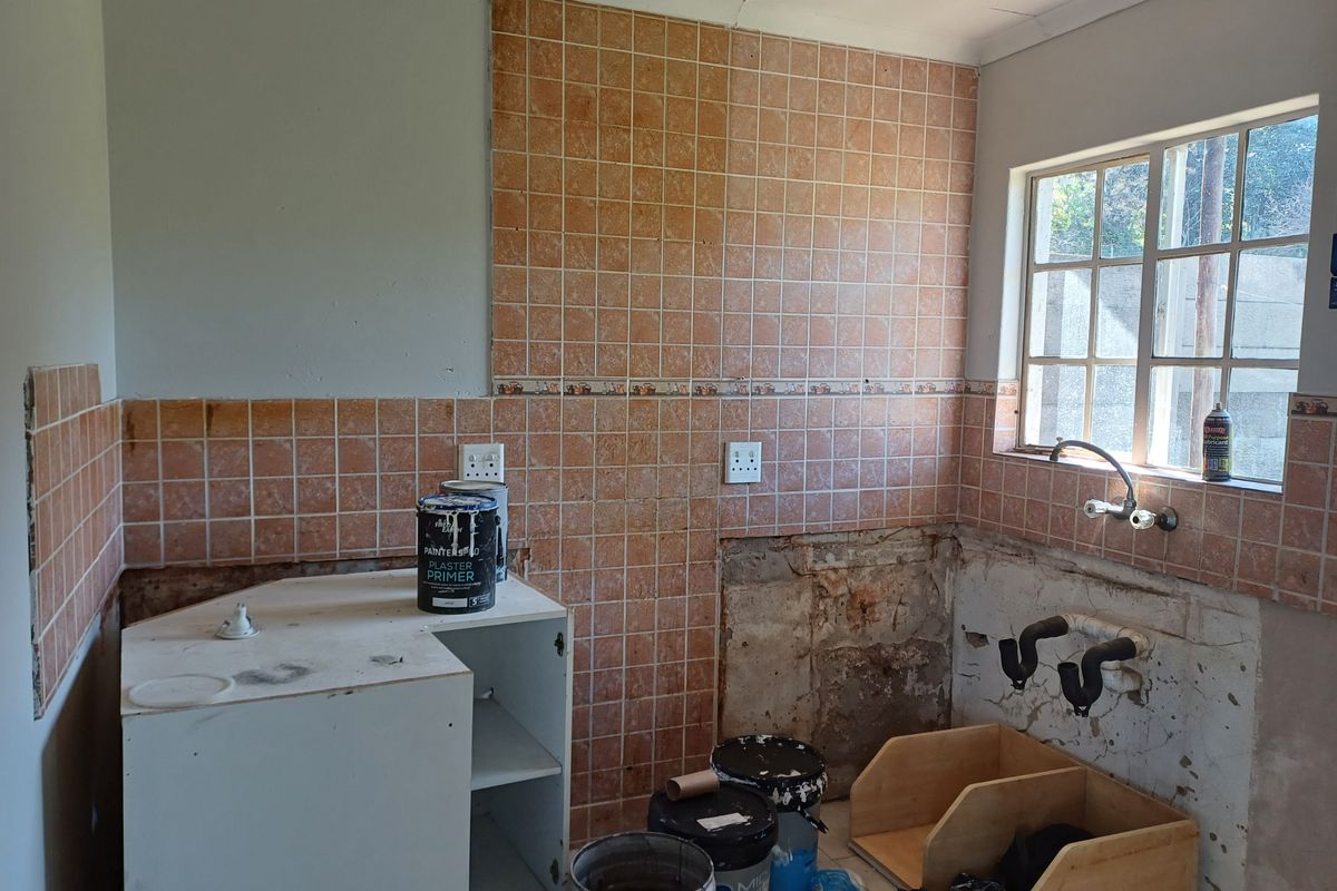 A kitchen space currently undergoing renovation, featuring tiled splashbacks, a window, and exposed plumbing connections ready for new cabinetry and fixtures.
