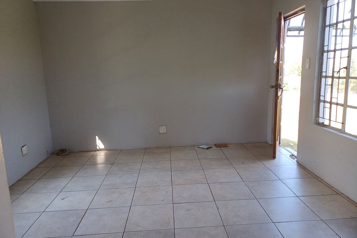 An empty living room featuring neutral tiled flooring, security bars on the windows, and direct access to the exterior.