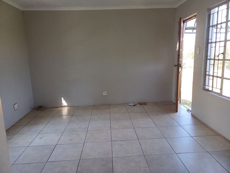 An empty living room featuring neutral tiled flooring, security bars on the windows, and direct access to the exterior.
