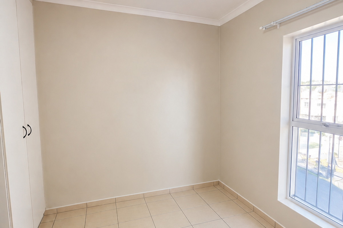 A clean, empty bedroom featuring neutral tiled flooring, a built-in wardrobe, and a window fitted with security bars.