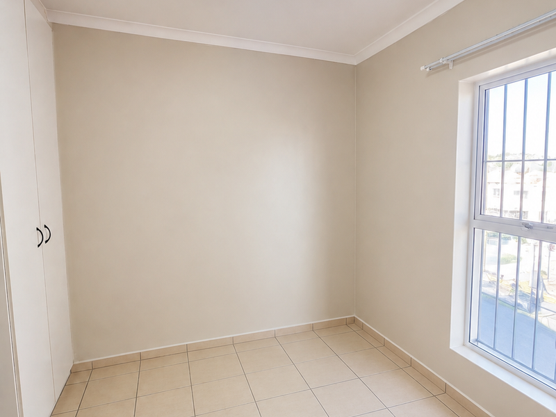 A clean, empty bedroom featuring neutral tiled flooring, a built-in wardrobe, and a window fitted with security bars.