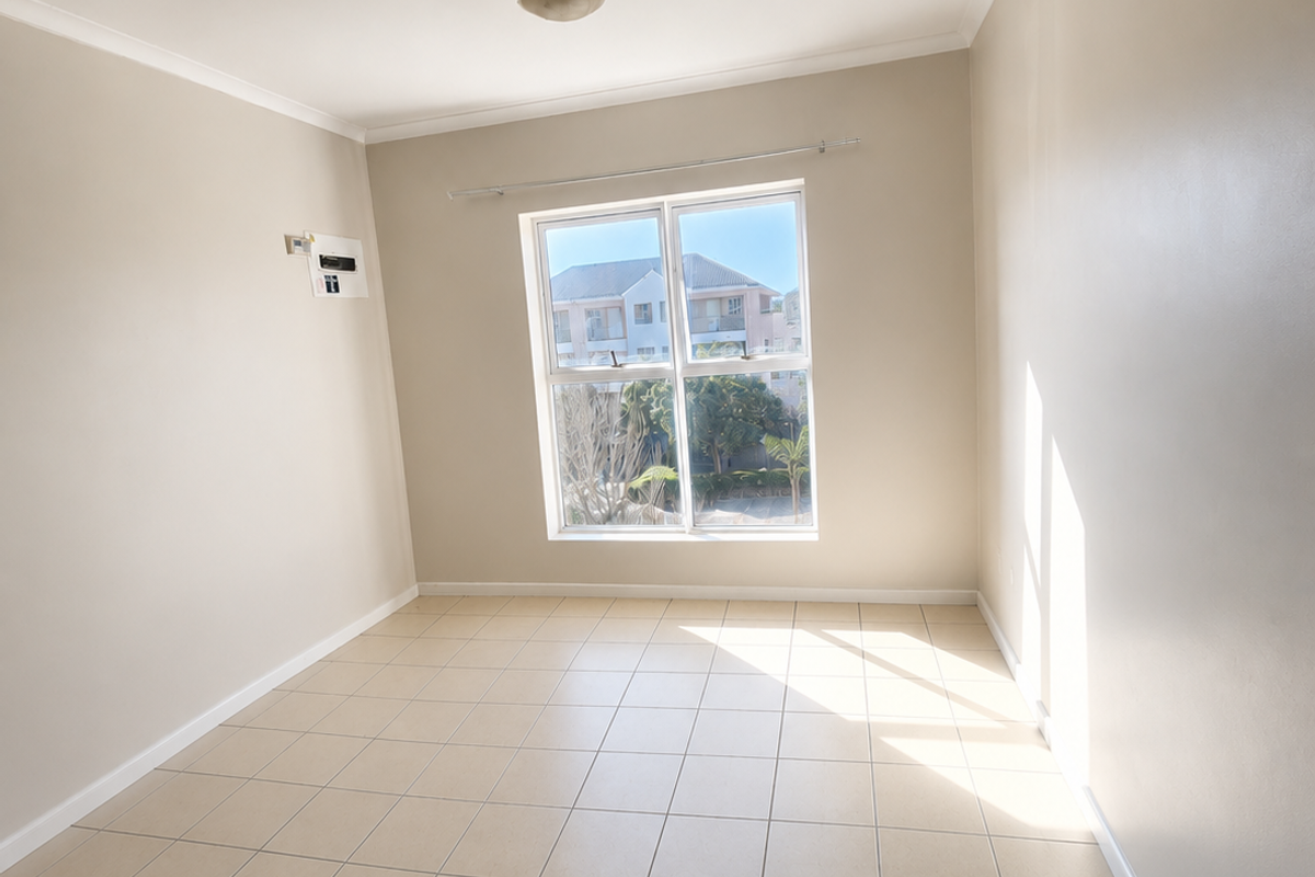 A bright, empty bedroom featuring neutral tiled flooring, a central ceiling light, and a window providing natural light and neighborhood views.